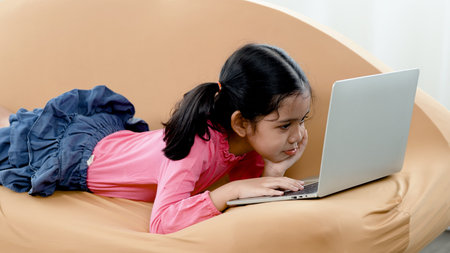 An adorable Asian girl wearing a pink shirt is happily sitting at her laptop notebook computer on the sofa in the living room of her home.の写真素材
