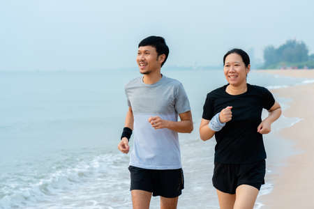 This young Asian couple, jogging in the cold wind together in the morning, the weather is nice and cool, the sea waves hit their feet while running, feel relaxed.の写真素材