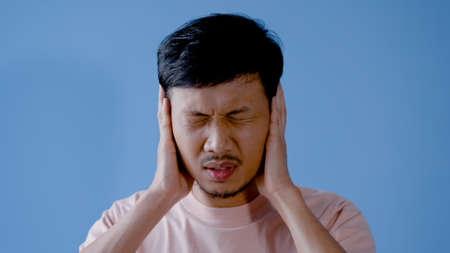 Asian man mustache,Half of the young man raised his hands to cover his ears , in frustration , because the surroundings were too noisy.Isolated indoor studio on blue background.の写真素材
