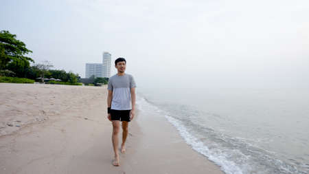 Young Asian man walking by the sea in the morning with good weather, clear skies, in the background with the morning sun rising, popular tourist attractionの写真素材