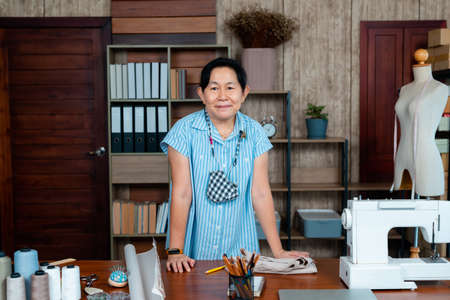 Professional small business owner partner in clothing design, close-up of Asian senior woman  standing at her desk full of sewing supplies, online shop business idea.の写真素材