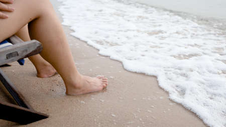 Close-up of sea waves crashing against the feet of a young Asian man who sits slowly working in a chair, feeling cool,work from home.の写真素材