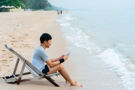 A handsome Asian man, chilling with his mobile phone by the sea in the morning, with cool air and clear skies, comfortably.work for, homeの写真素材