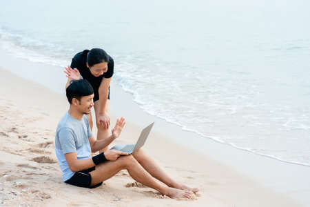 Good-looking Asian guy, video chatting with his colleague on his laptop computer, and another friend came to say hi, sat by the sea in a good mood,work from home.の写真素材