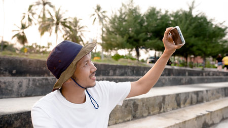 Nice looking Asian man wearing his favorite hat, sitting on a chair and taking a selfie on the...