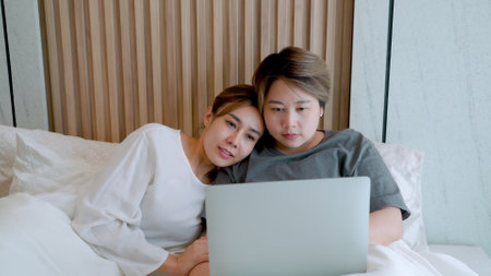 Two Asian women, who are lovers, They both were looking at something on their laptop computers, happy laughing expressions, on the white bed in the room, the love, and friendship of LGBTQ couplesの写真素材