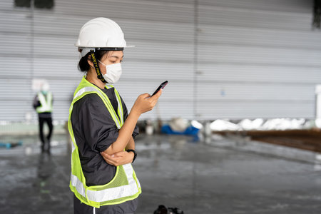 An Asian female engineer wearing a green reflective vest wears a protective hard hat. A foreman...