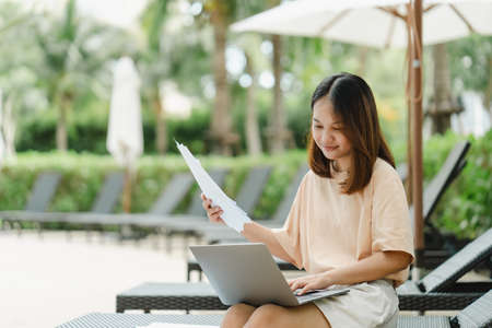 close-up A woman working at the swimming pool with her laptop computer holding paperwork in her hand. It is imperative for young Asian female tourists to sit and work on vacations at the hotel.の写真素材