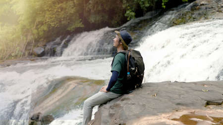 Asian young man, sitting with his backpack, sitting on the edge of a large rock, enjoying the wind and watching nature by the waterfall, in the daytime with a pleasant breeze.の写真素材