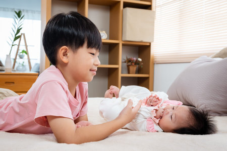 A 9-year-old boy is playing in bed with his 3-month-old newborn sister. An Asian boy is taking care of his sister in bed while waiting for his mother to prepare milk for his sister to drink.の写真素材