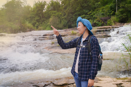 Asian female trekker with travel gear, backpack and crutches stands by the waterfall in the forest, admiring the beauty of nature on weekends, wishing to be happy on a clear day.の写真素材