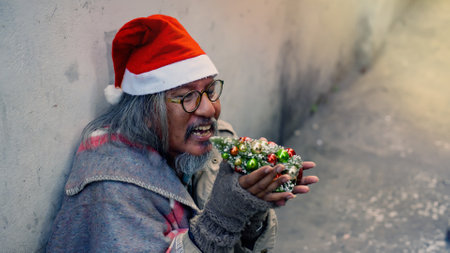 An old homeless Asian man wearing a red Christmas hat holding a small Christmas tree is showing signs of a mental breakdown. The madman sat alone on the roadside laughing and smiling.の写真素材