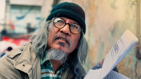 An old Asian man in glasses and a black hat, dressed in a dirty, homeless man, is reading the newspaper in search of work and help. Homeless people who do not have home to sleep on streetsの写真素材