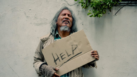 A homeless, long-haired Asian man stands desperately in a lonely alley wall and in his hand a sign asking for help because he has no work and no food to buy because he has no money.の写真素材