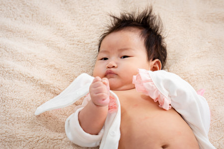 Close-up of a 3-month-old Asian newborn lounging in bed with a happy smile on her face and wriggling with her mother's teasing, covering her mouth with her hands.の写真素材