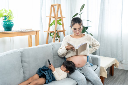 cute little daughter lying on mom's lap playing mobile phone and a pregnant Asian single mom reading a book comfortably on her living room sofa withの写真素材