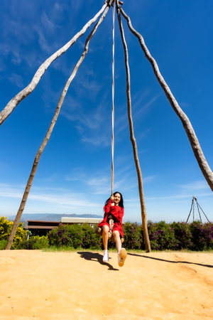 Asian teenage girl with long hair wearing red sweater playing with a wooden swing in the weekend tourist area. The joy of traveling in the countrysideの写真素材