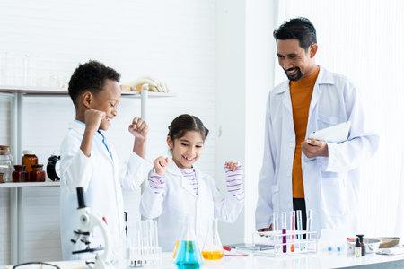 .In chemistry classroom with many laboratory tools on shelves and table. A male teacher in white lab coat confirms experimental result from a young African boy and little Asian girl with joy together.の写真素材