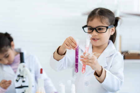 In chemistry classroom with many laboratory tools. Two little Asian girl in white lab coat help each other for experiment. A girl with glasses pick up orange and violet solution test tubes carefully.の写真素材
