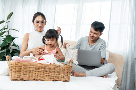 Beautiful pregnant Asian mother, sitting folding clothes with tiny daughter, on bed, behind is husband, sitting at work, on laptop computer, sat watched wife and children accidentally smiled.の写真素材