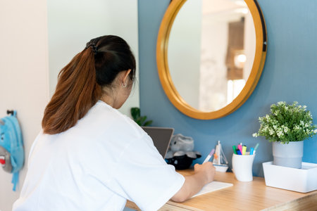 Asian girl teenager with long hair wearing white t-shirt sitting upright doing homework in bedroom When studying at home, you have to sit and study online and on a computer.の写真素材