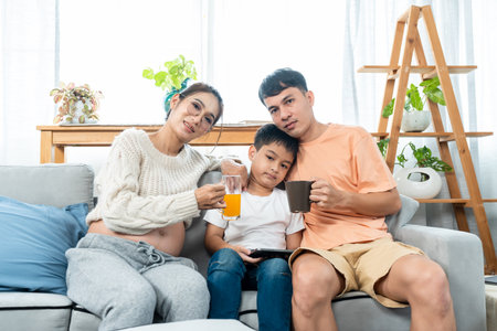 In a cozy living room, an Asian family spend their free time together. A pregnant mother holding a glass of orange juice and sitting with her son with his tablet and husband holding a cup of coffee.の写真素材