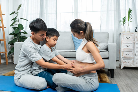 Beautiful Asian mom, sitting on carpet in bedroom, father and son sit across from mother, father took both of sons' hands, gently stroking mother's big belly, touching sister in mother's womb.の写真素材