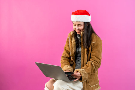 Beautiful Asian girl in long-sleeved shirt wearing Christmas hat, puts her laptop on her leg, doing her assignments, expression looking serious at work, Isolated indoor studio on pink background.の写真素材
