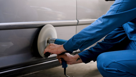 Close-up hand male mechanic wear mechanic's uniform for work safety, sit down use car polisher scrub slowly hold machine firmly make car's surface smooth before going down use new paint spray down.の写真素材