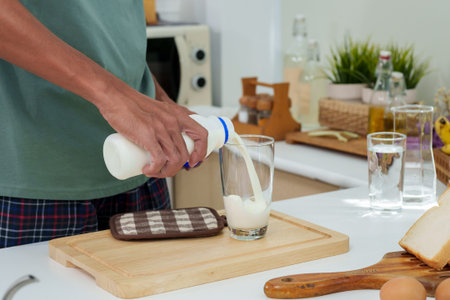 Close-up Asian man's hand standing in kitchen in morning, man opened cap bottle plain cow's milk come out pour it into glass that sits on table with mango hair fresh chicken eggs.の写真素材