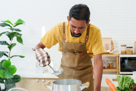 Asian man with mustaches Clad in an apron preparing dinner, cooking soup on the stove. Open the lid of the pot, smoke from hot boil comes out. It's in kitchen. On days when have to stay home aloneの写真素材