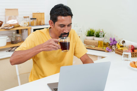 Close-up Asian man's eyes sitting on laptop computer, in living room at home, sit watching fun clips in online apps, on lazy days off from work, holding coffee mug in hand drink coffee.の写真素材