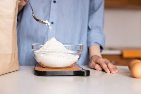 Set of ingredients for cooking according to the recipe: flour, eggs. Woman puts a spoon of flour in bowl on the scales, as much as necessary for the recipe, hands in the frame, Cooking at home. Seriesの写真素材