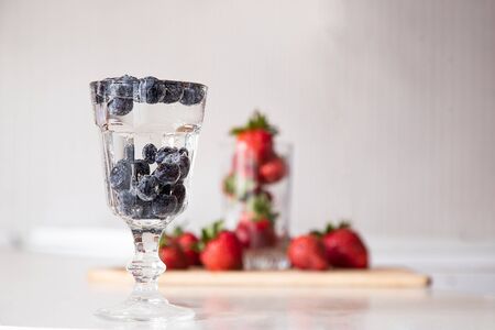 A glass of water and blueberries on the kitchen table. Beautiful air bubbles and fresh berries in the water. On the background is a glass of strawberries. Berries on the table.の写真素材