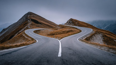 Two-lane asphalt road winds through a mountainous landscape, diverging at a fork in the road, with dramatic autumnal colors and a misty sky above.の素材