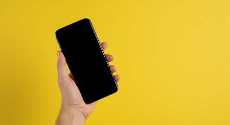 Female hand holding black smartphone with blank screen on yellow background with copy space.の素材