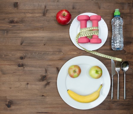 Healthy lifestyle concept. colored Apples measuring tape dumbbells banana look like face sport  water on  wooden tableの写真素材
