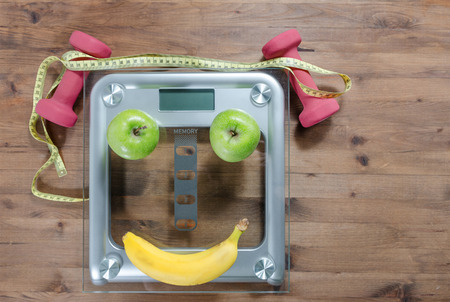 Healthy lifestyle concept. scales colored Apples measuring tape dumbbells banana look like face on wooden tableの写真素材