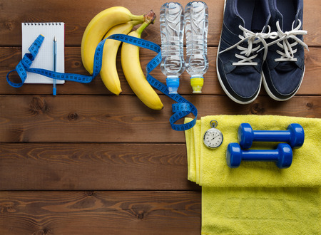 Fitness concept with sneakers dumbbells stopwatch measure tape towel bottle of water and bananas on wooden table backgroundの写真素材
