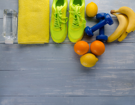 Fitness concept with sneakers dumbbells towel bottle of water bananas oranges and lemons on blue wooden table background.の写真素材
