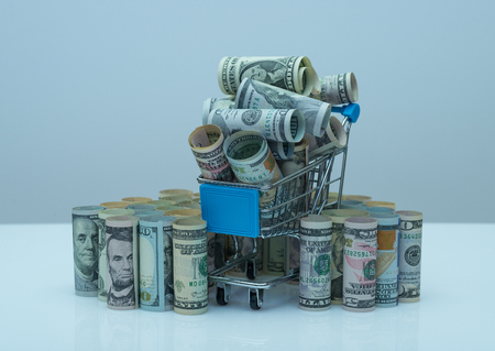 US dollars rolls and shopping cart full packs of bills on colored background with reflection. Creative business and financial concept with usa dollar banknotesの写真素材