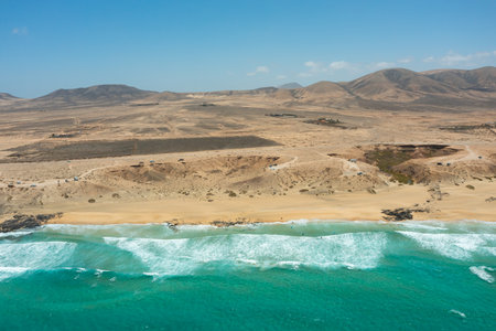 Spectacular aerial view of Playa del Castillo in Caleta de Fuste, Fuerteventura, Canary Islands. The photo showcases the vibrant turquoise waters, golden sand, and historic Castillo de San Buenaventura, all bathed in sunlight. Kitesurfers glide across the waves, adding dynamic energy to this idyllic coastal scene. A perfect blend of natural beauty, water sports, and sunny island atmosphere.の写真素材