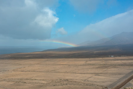 A stunning landscape of Fuerteventura's dry plains leading towards the Atlantic coast, with a dirt road under a vibrant rainbow after the rain.の写真素材
