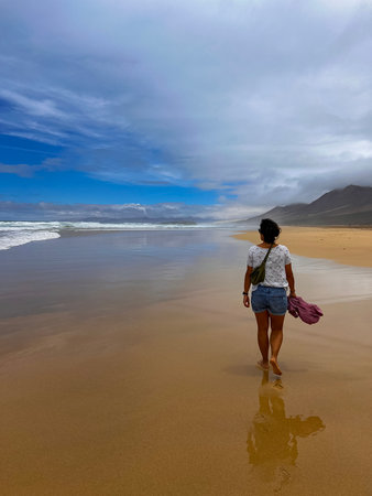 Traveler enjoying a sunny yet cloudy day at Cofete Beach, Fuerteventura. A young woman walks by the ocean with a light backpack, surrounded by natural beauty and freedom.の写真素材