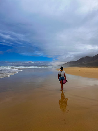 Young woman on vacation walking along the wild Cofete Beach in Fuerteventura, Canary Islands. She carries a small crossbody backpack under a sky of sun and clouds.の写真素材