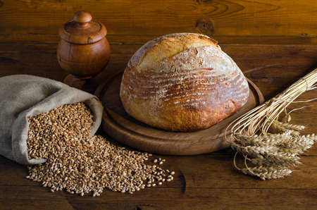 Freshly baked bread, spikelets and small bag with wheat grains on a wooden table. Rustic style.の写真素材