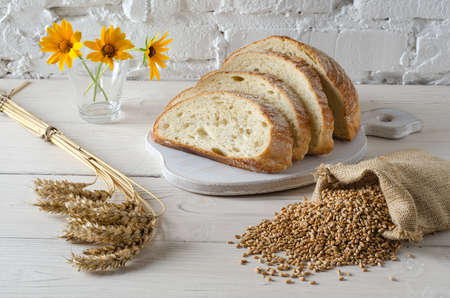 Sliced bread on cutting board and small bag with wheat grain on white wooden table. Rustic style.