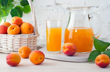 Apricot juice in glass and decanter with ripe apricots on wooden table on background of white brick wall with green leaves.の写真素材