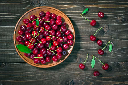 Red cherries in a plate, green leaves on a dark wooden table. Rustic style, top view.の写真素材