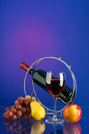 Glass with red wine, bottle of wine on the stand, brush of grapes and fruits on bright multicolor background with reflection. Copy-space.の写真素材